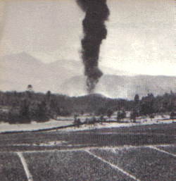 a column of black smoke rises out of a hole in a corn field